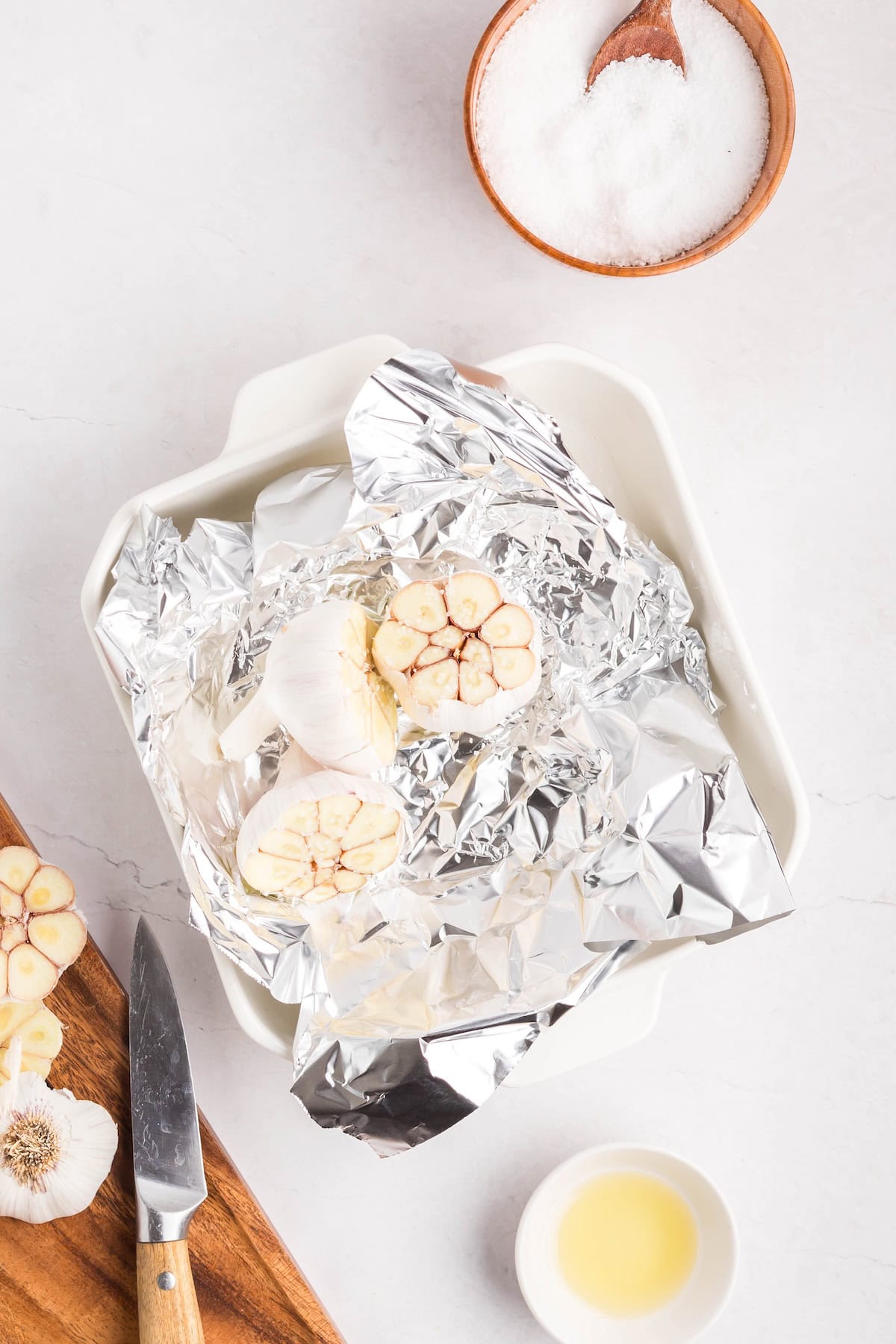 A baking dish with halved garlic bulbs ready for roasted garlic rests on aluminum foil, alongside a knife, a bowl of salt, and a small bowl of oil, all arranged on a white surface.