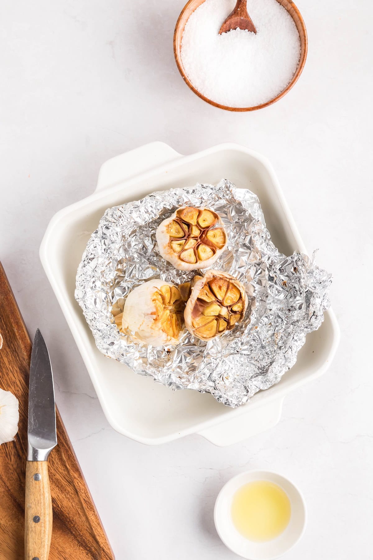A head of roasted garlic cut in half sits on aluminum foil in a white baking dish, with a knife, a bowl of salt, and a small cup of oil nearby—perfect for spreading this flavorful roasted garlic on your favorite dishes.