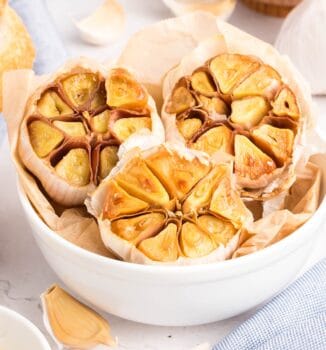A white bowl filled with roasted garlic—halved heads glistening on parchment paper—sits amid raw garlic cloves on a light surface.