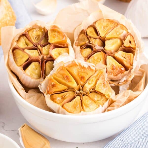 A white bowl filled with roasted garlic—halved heads glistening on parchment paper—sits amid raw garlic cloves on a light surface.