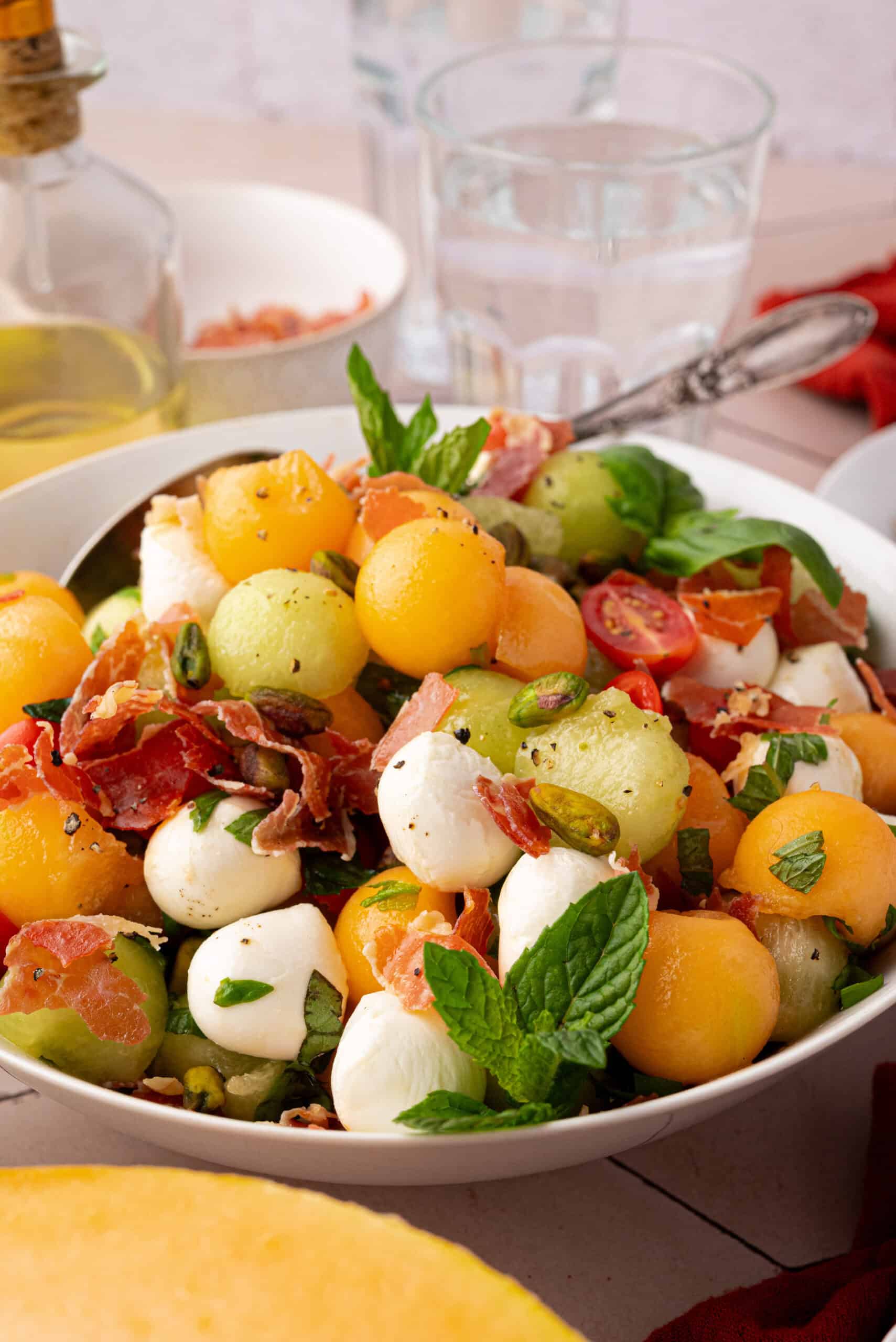 A Summer Fruit Caprese Salad featuring melon balls, mozzarella, prosciutto, cherry tomatoes, basil, and pistachios is served in a white bowl with a spoon.