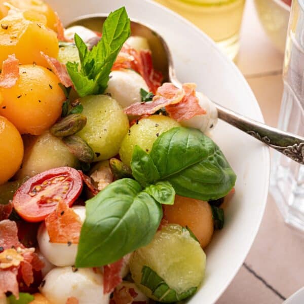 A close-up of a Summer Fruit Caprese Salad with melon balls, cherry tomatoes, mozzarella, basil leaves, prosciutto, pistachios, and a spoon in a white bowl.