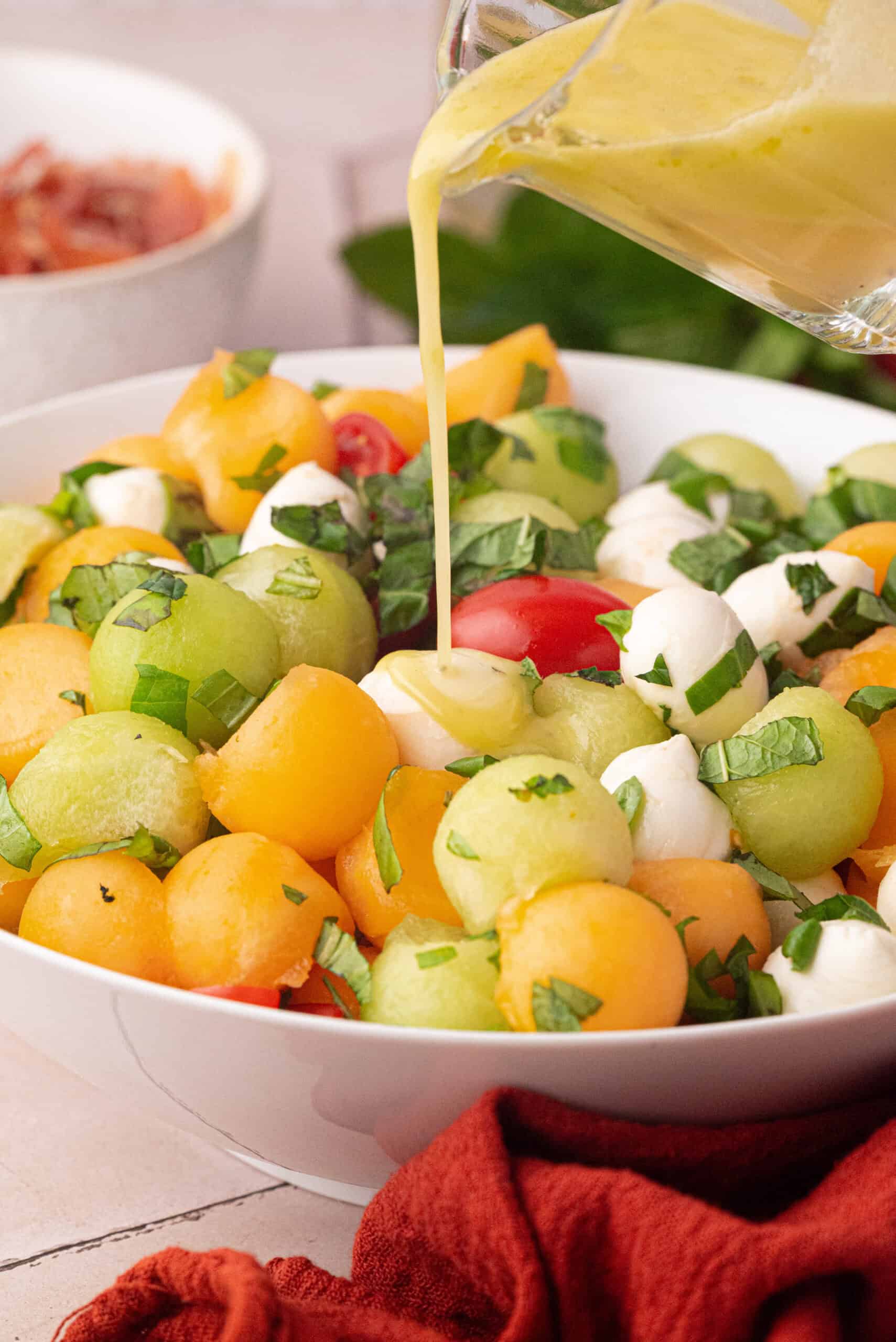 A refreshing Summer Fruit Caprese Salad features melon balls, cherry tomatoes, and mozzarella balls, garnished with chopped herbs as a vibrant yellow dressing is poured over the bowl.