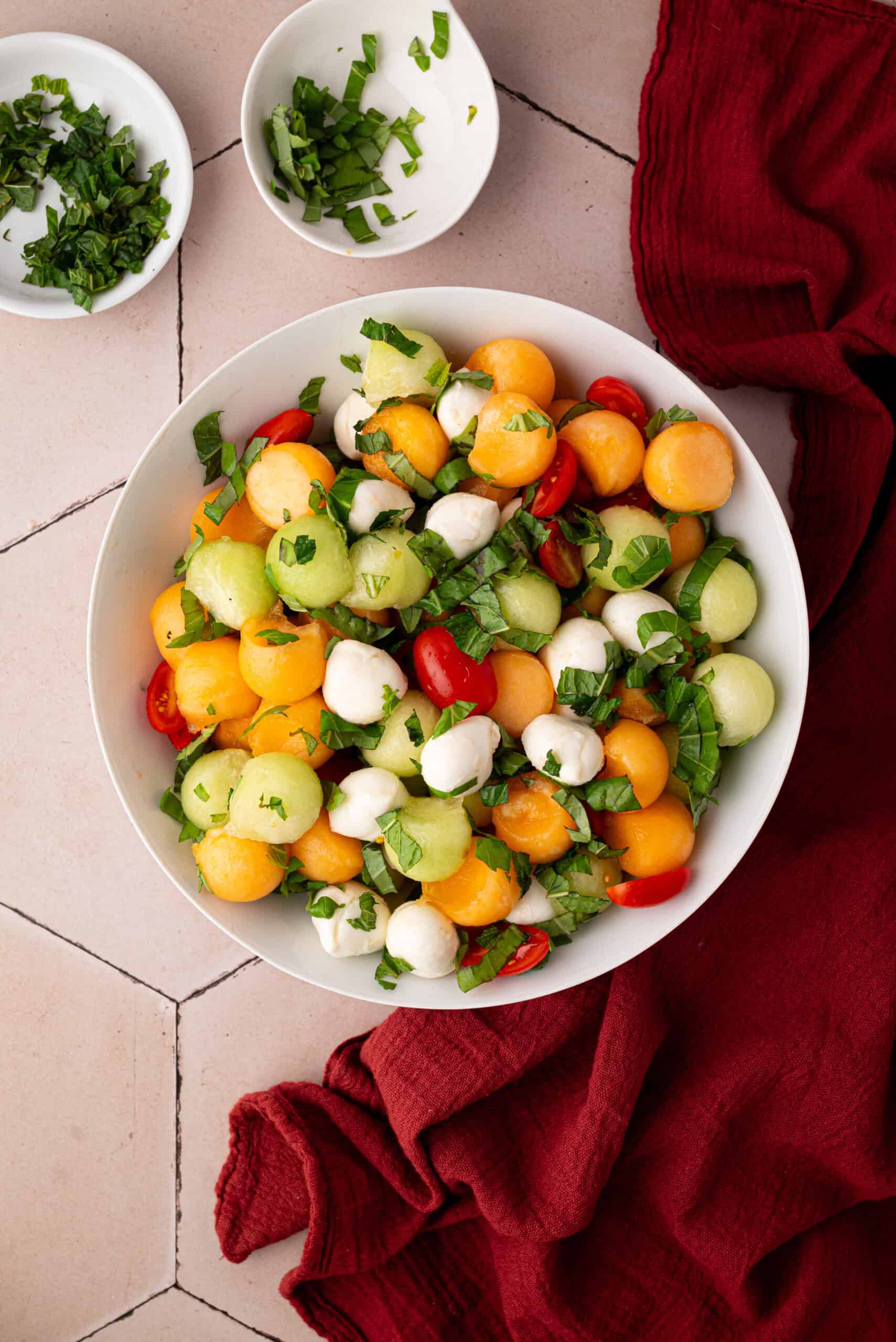 A bowl of Summer Salad with melon balls, cherry tomatoes, mozzarella balls, and chopped basil sits on a tiled surface next to two small bowls of herbs and a red cloth.