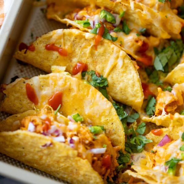 Baked Chicken Tacos with a crispy hard shell are filled with shredded chicken, melted cheese, chopped onions, cilantro, and drizzled with red sauce, all arranged closely together on a tray.