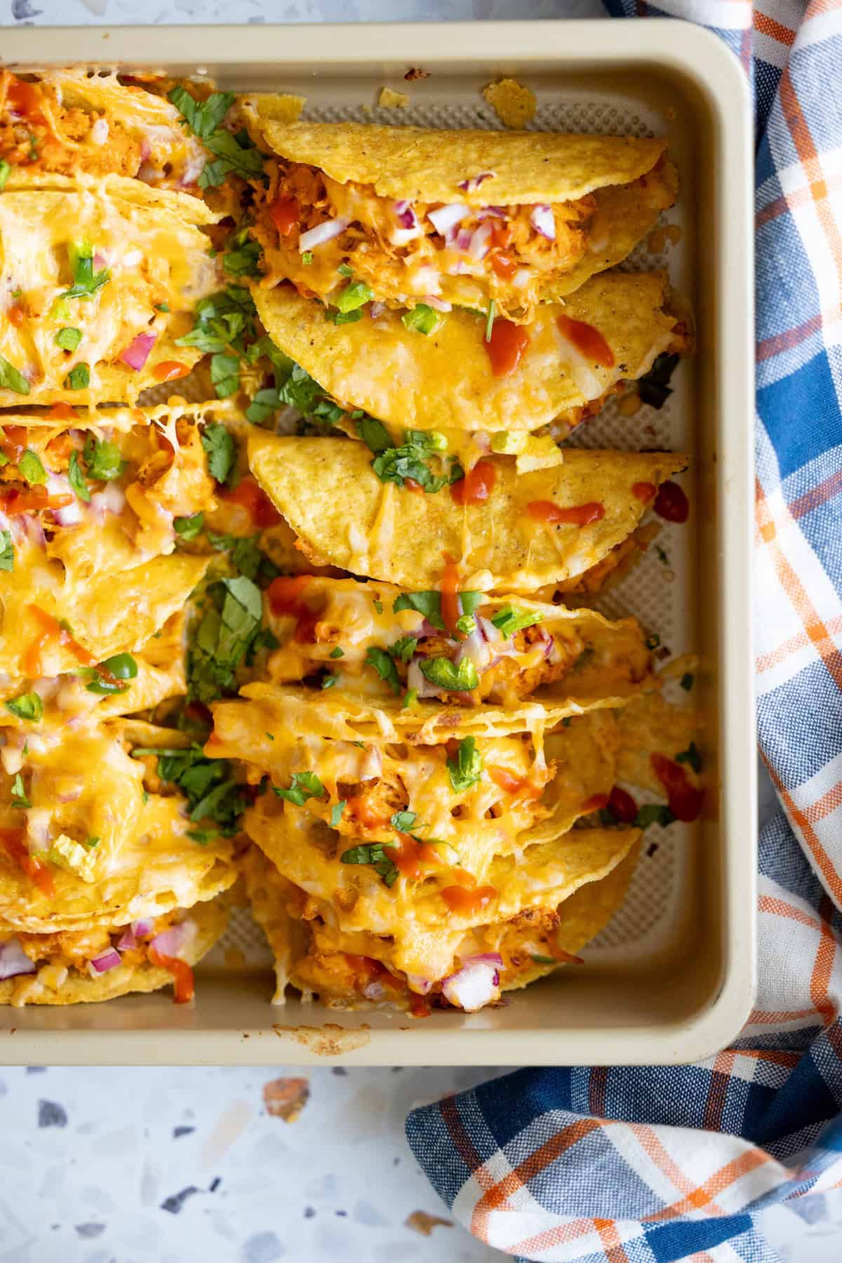 A baking tray filled with baked chicken tacos topped with melted cheese, diced red onions, chopped green herbs, and sauce, next to a blue and orange plaid cloth.