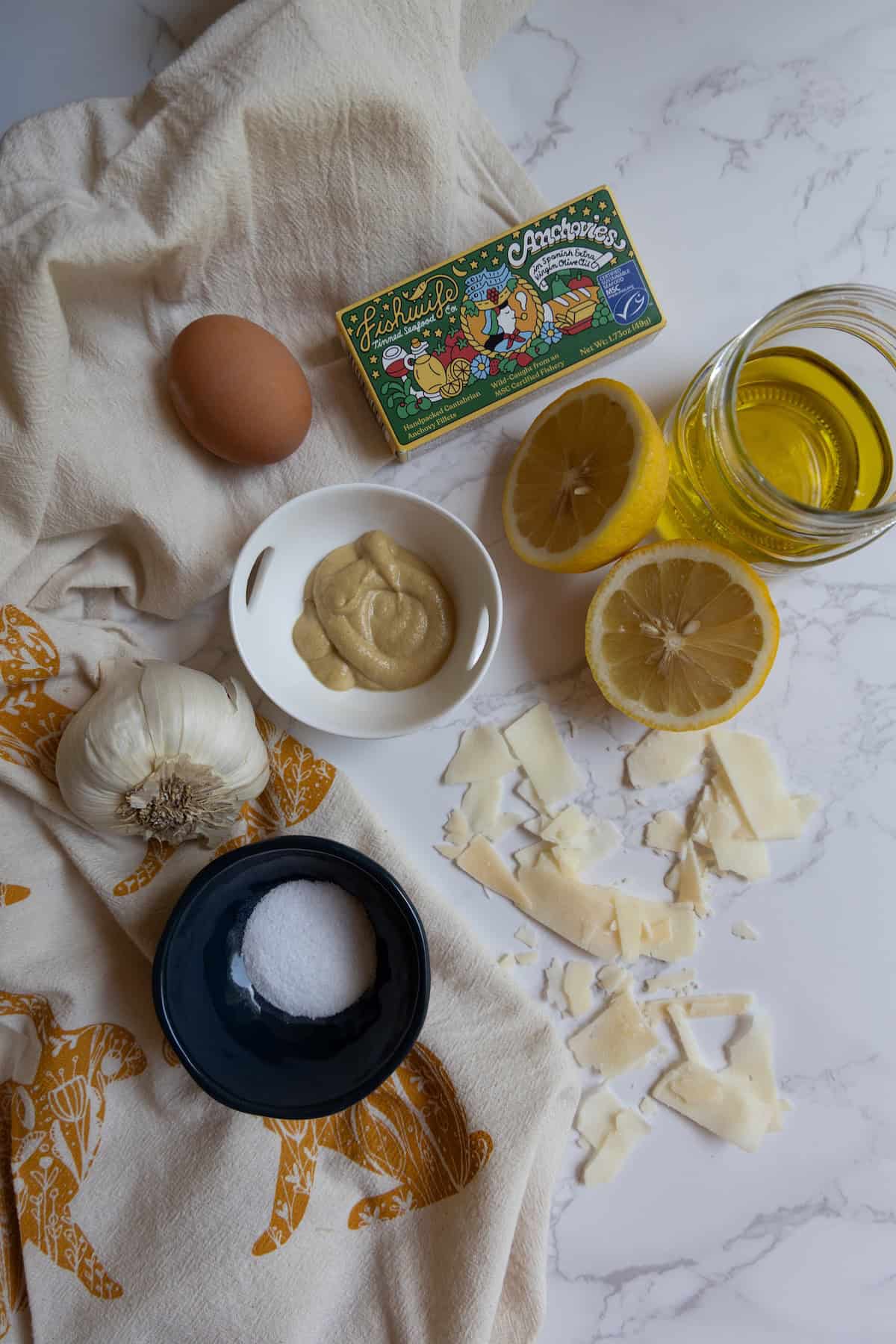 Ingredients for a caesar dressing recipe arranged on a white surface: an egg, mustard, lemon halves, olive oil, garlic, salt, cheese shavings, and a box of anchovies.