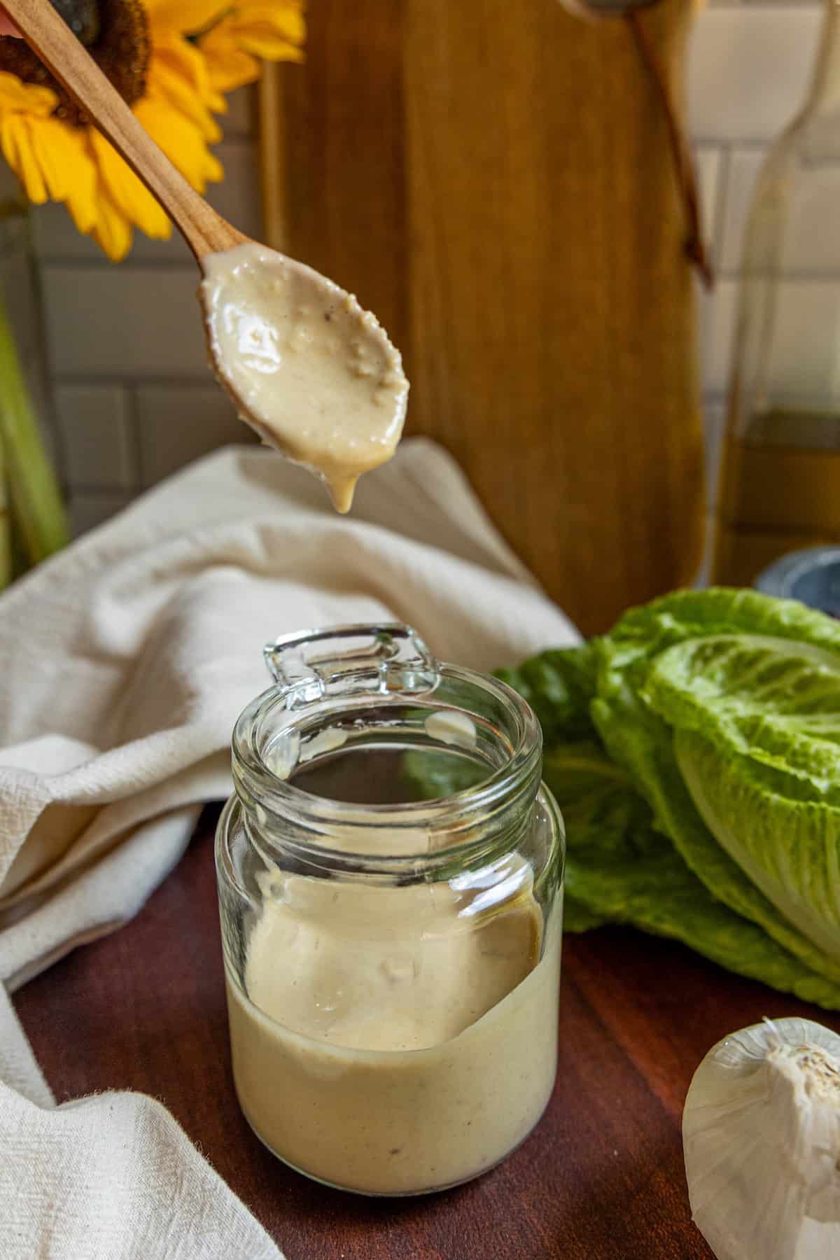 A wooden spoon with creamy caesar dressing hovers over an open glass jar; lettuce, garlic, and a cloth rest nearby on the wooden surface.