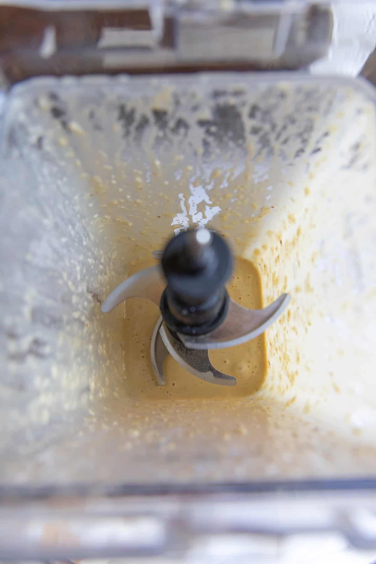 Top-down view of a blender containing a creamy, light yellow caesar dressing mixture with remnants stuck to the sides and the metal blades visible in the center.
