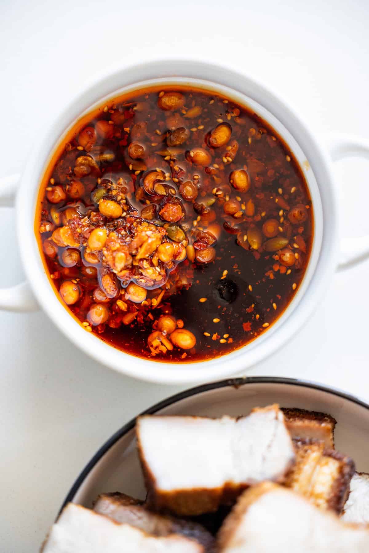 A white bowl filled with chili oil containing chili flakes and seeds sits next to a bowl of Sticky Asian Pork Belly, featuring perfectly sliced roasted pieces.