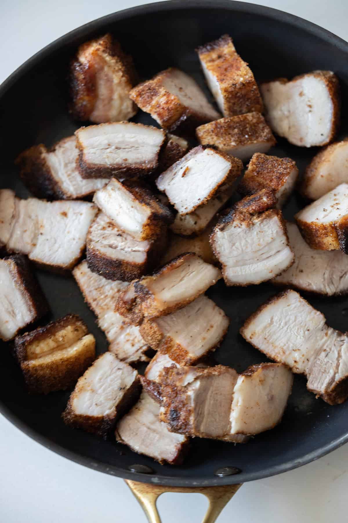 Sliced pieces of Sticky Pork Belly with crispy, browned edges are arranged in a black skillet with a gold handle.