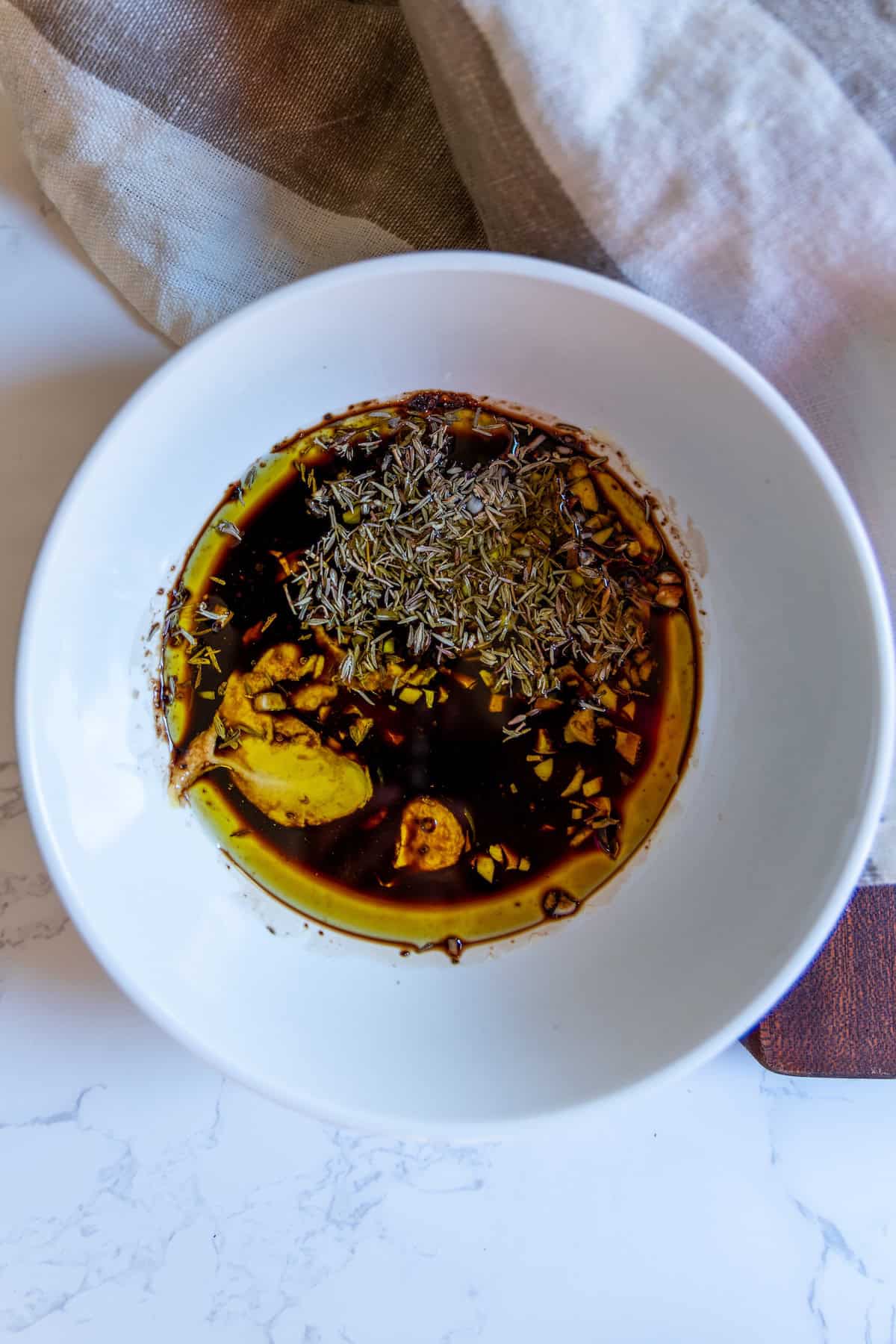 A white bowl containing a mixture of soy sauce, olive oil, garlic slices, dried herbs, and spices on a marble surface—perfect as a flavorful dressing for the best cobb salad.