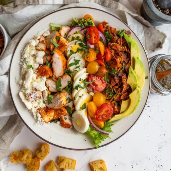 A bowl of the best Cobb salad with sliced grilled chicken, hard-boiled eggs, cherry tomatoes, red onion, bacon, avocado, crumbled cheese, and fresh greens.