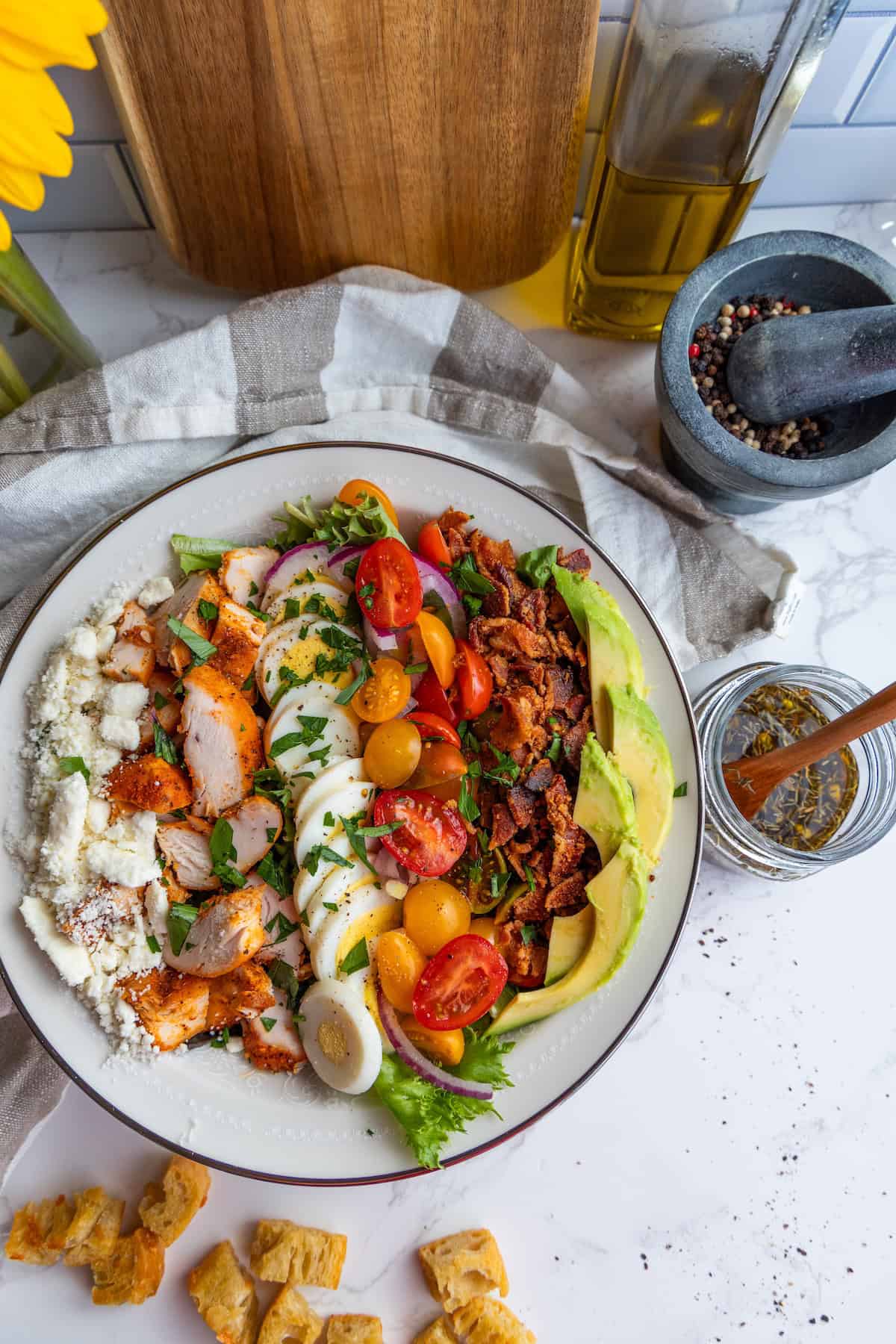 Enjoy the best Cobb salad: a plate filled with grilled chicken, boiled eggs, cherry tomatoes, avocado, bacon, red onion, feta cheese, and mixed greens. Dressing, croutons, and seasonings are served on the side.
