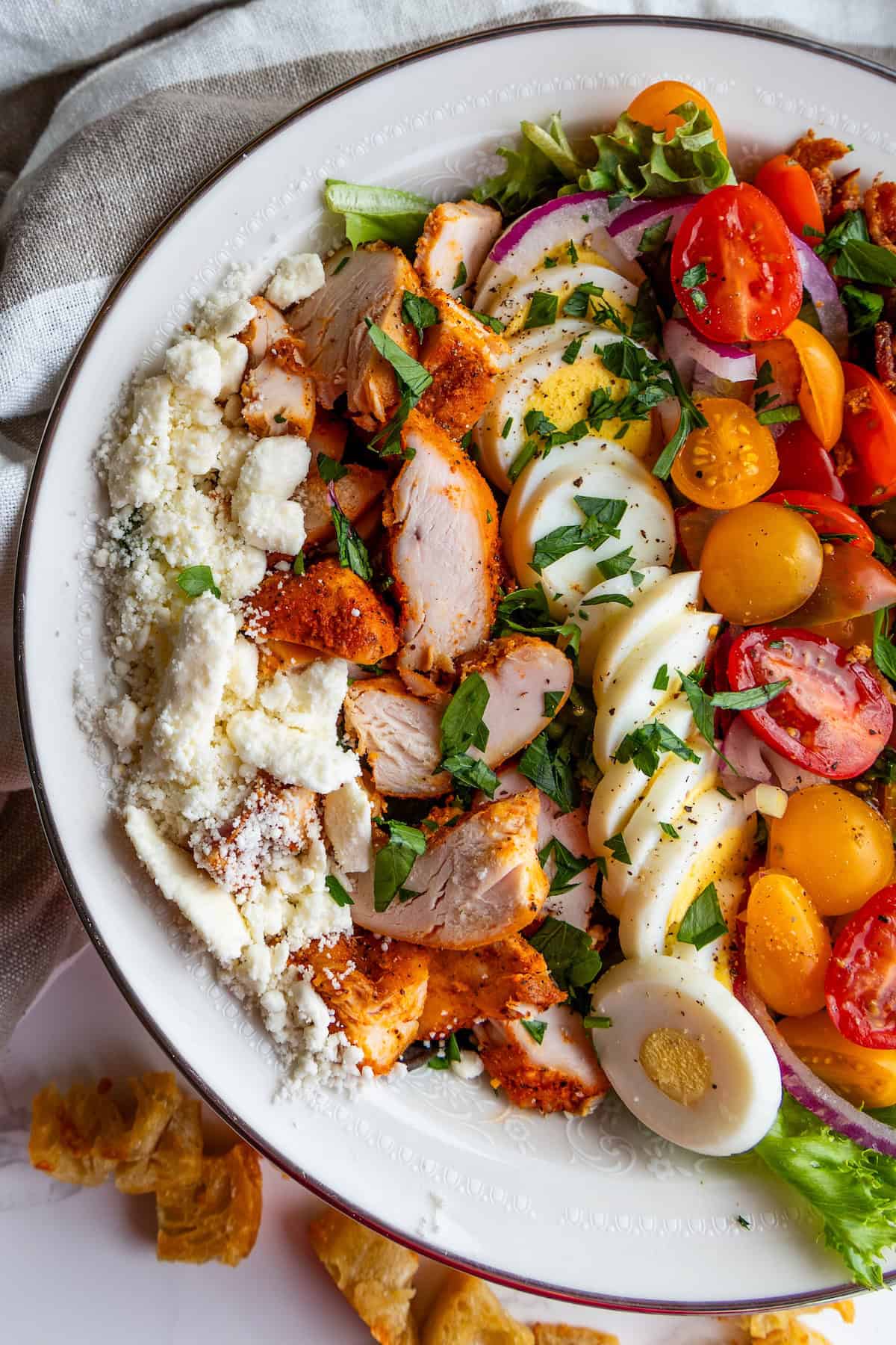 A plate of the best Cobb salad with sliced grilled chicken, hard-boiled eggs, cherry tomatoes, red onions, lettuce, and crumbled cheese, all garnished with chopped parsley.