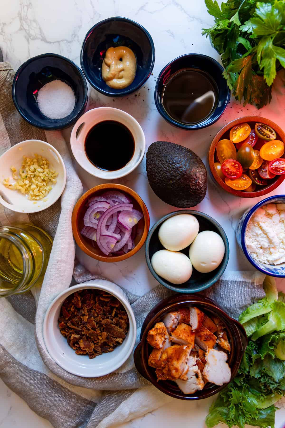 A flat lay of the best Cobb salad ingredients, featuring eggs, avocado, grilled chicken, bacon, red onion, grape tomatoes, cheese, sauces, oil, salt, minced garlic, and fresh greens on a marble surface.