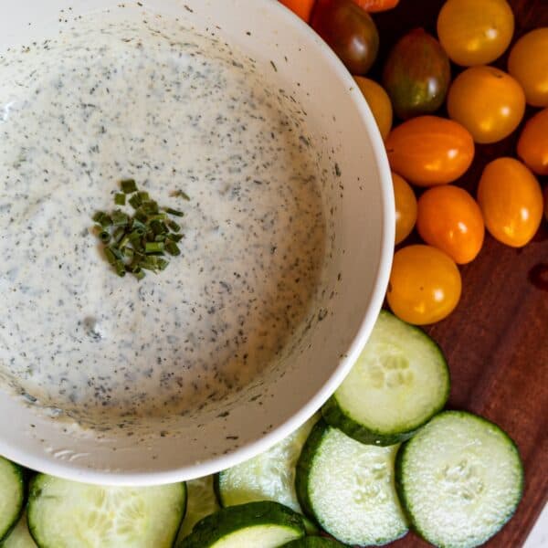 A bowl of creamy homemade ranch dressing dip garnished with chopped chives is surrounded by sliced cucumbers and yellow cherry tomatoes on a wooden surface.