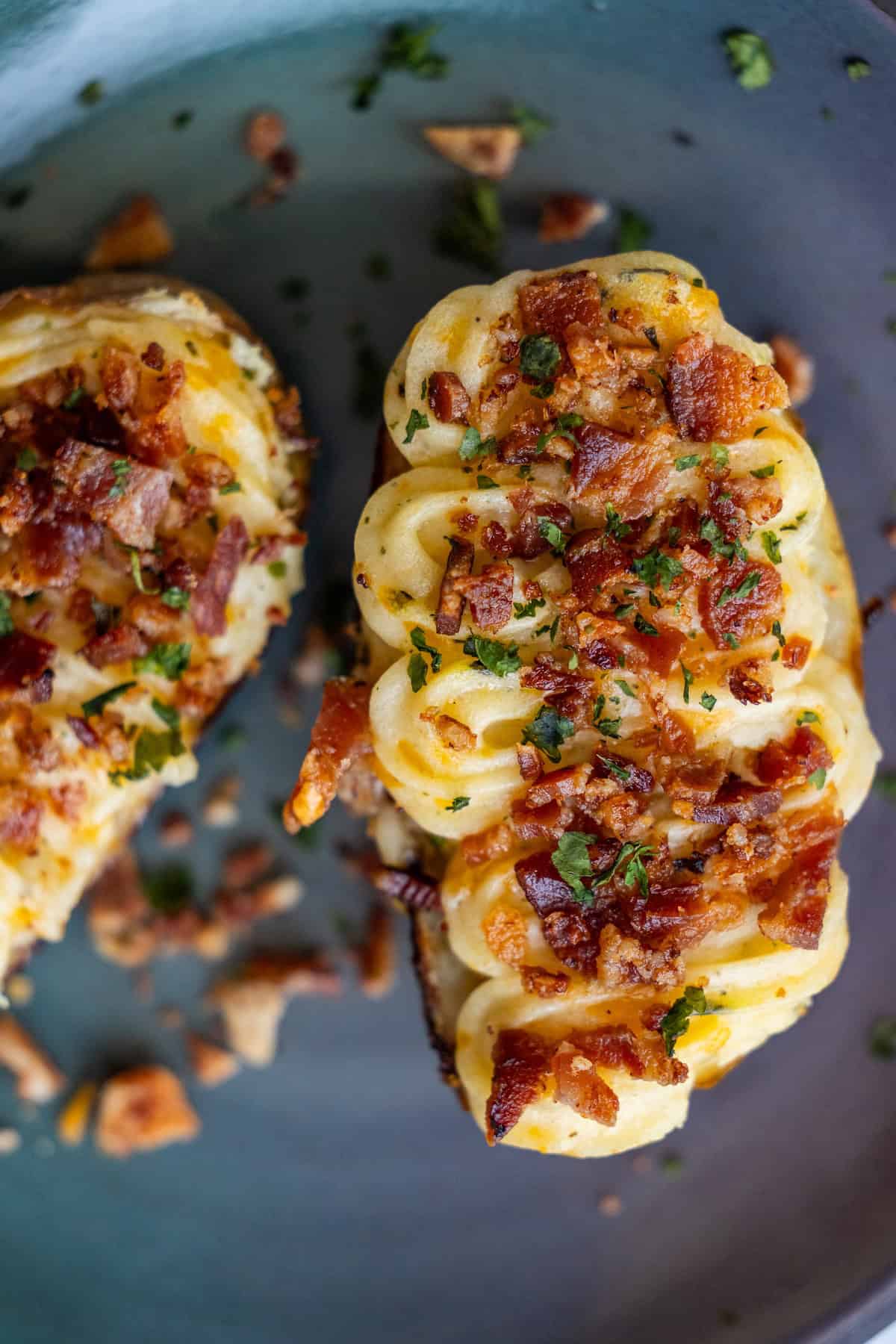 Close-up of a twice-baked potato topped with crumbled bacon, chopped herbs, and piped mashed potatoes on a plate.