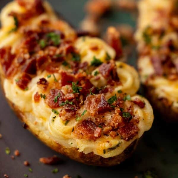 A close-up of Loaded Twice Baked Potatoes topped with melted cheese, crumbled bacon, and chopped herbs on a dark surface.