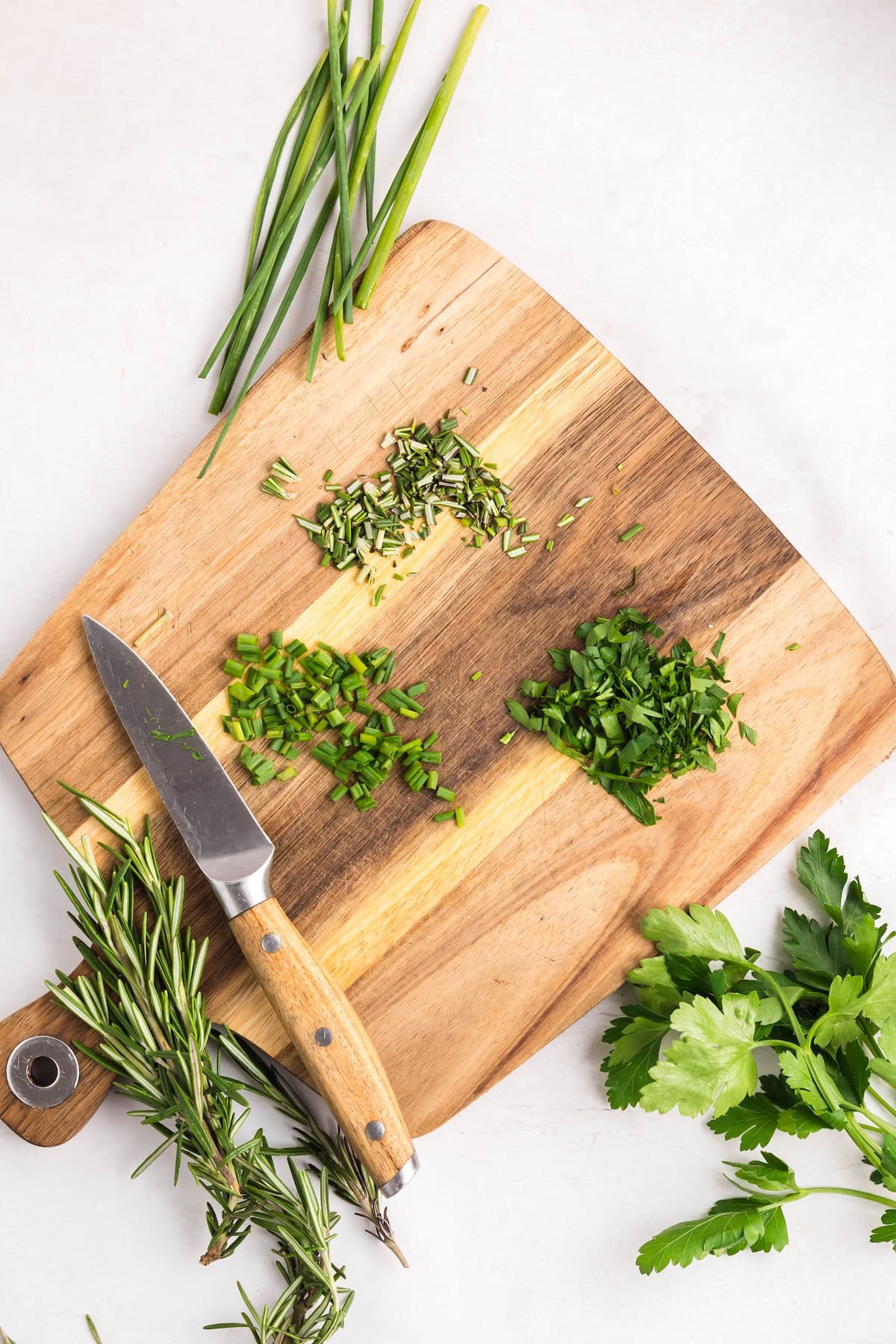 A wooden cutting board with chopped rosemary, chives, parsley, and hints of roasted garlic butter, alongside a knife, whole herbs, and green onions arranged on a white surface.