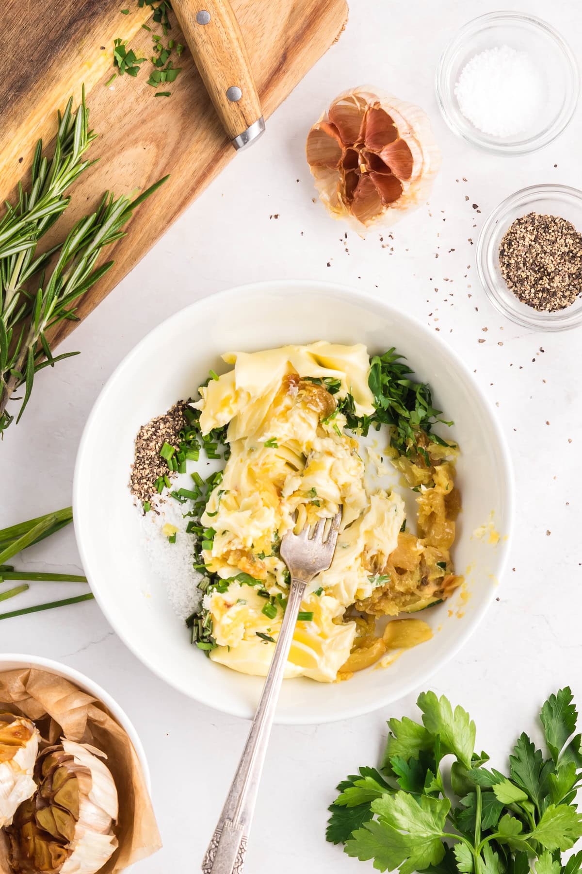 A bowl with softened butter, chopped herbs, roasted garlic, salt, and pepper is being mixed with a fork to create creamy Roasted Garlic Butter; fresh herbs and seasonings are arranged nearby on a white surface.