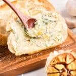 A slice of bread on a wooden board is being spread with rich roasted garlic butter using a wooden spoon, with a head of roasted garlic nearby.
