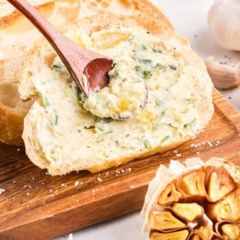 A slice of bread on a wooden board is spread with roasted garlic butter using a wooden spoon; a halved roasted garlic bulb is nearby.