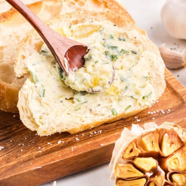 A slice of bread on a wooden board is spread with roasted garlic butter using a wooden spoon; a halved roasted garlic bulb is nearby.