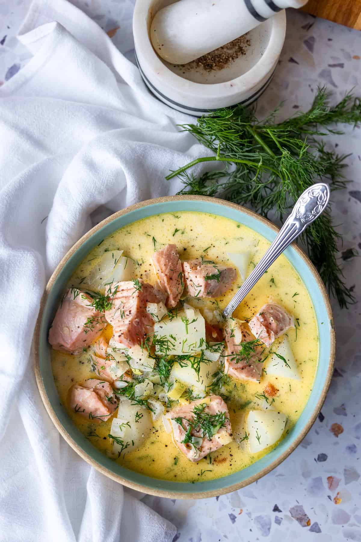 A bowl of creamy Finnish salmon chowder, or lohikeitto, with chunks of salmon, potatoes, and fresh dill sits next to a white napkin, mortar and pestle, and sprigs of dill.