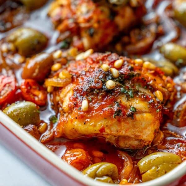 Baked harissa chicken thighs topped with herbs and pine nuts, surrounded by smashed olives and tomatoes in a vibrant red sauce.
