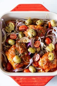 Baking dish with harissa chicken thighs, halved cherry tomatoes, sliced red onions, and smashed olives, ready to be cooked.