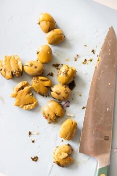 Green olives, some whole and some smashed olives, are scattered on a white cutting board next to a large kitchen knife.