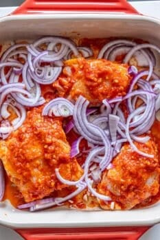 Raw chicken pieces coated in red sauce and topped with sliced red onions in a rectangular baking dish—perfect for making flavorful Harissa Chicken Thighs.