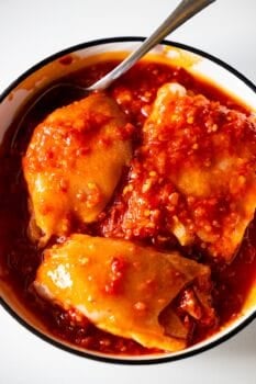 A bowl with three pieces of spicy chicken covered in a thick, red chili sauce, served with smashed olives and a spoon resting on the side.