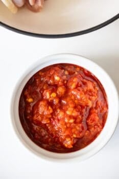A white bowl filled with chunky red chili paste, perfect for serving alongside spicy chicken thighs, is placed on a white surface.