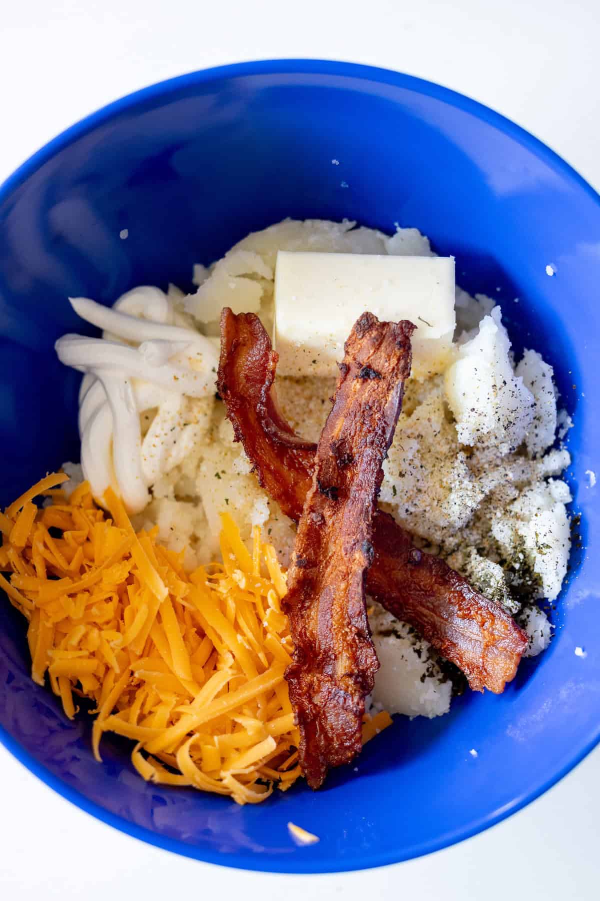 A blue bowl contains mashed potatoes, two strips of cooked bacon, shredded cheddar cheese, a pat of butter, mayonnaise, and seasonings—perfect for recreating a classic twice baked potatoes dish.