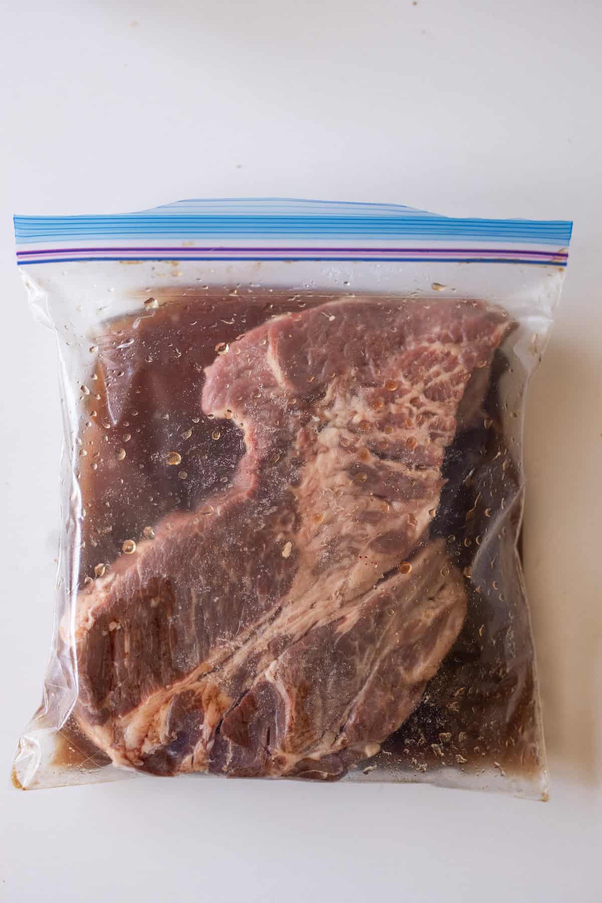 A large piece of raw meat marinating in a sealed plastic zip-top bag with a dark liquid, placed on a white surface.