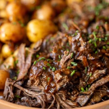 Close-up of shredded pot roast beef with gravy, garnished with chopped herbs, served alongside small roasted potatoes.