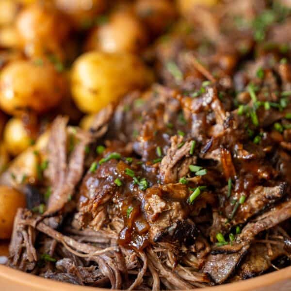 Close-up of shredded pot roast beef with gravy, garnished with chopped herbs, served alongside small roasted potatoes.