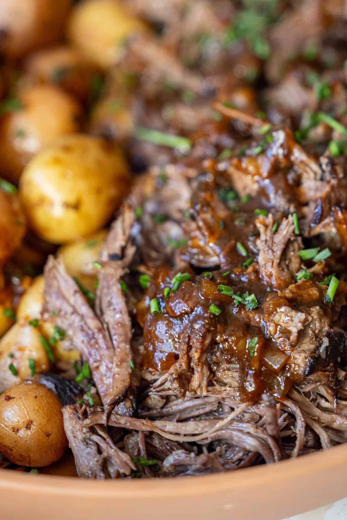 Close-up of shredded beef with brown sauce, garnished with chopped herbs, next to cooked baby potatoes.