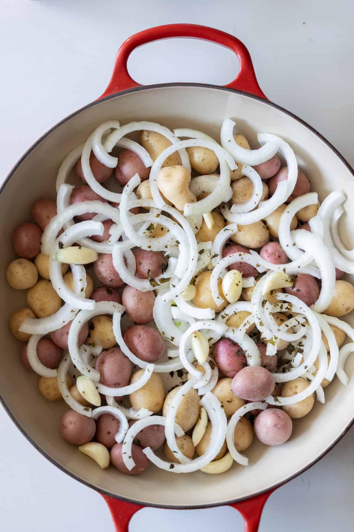 A red Dutch oven filled with halved baby potatoes, sliced onions, whole garlic cloves, and herbs, ready for cooking.