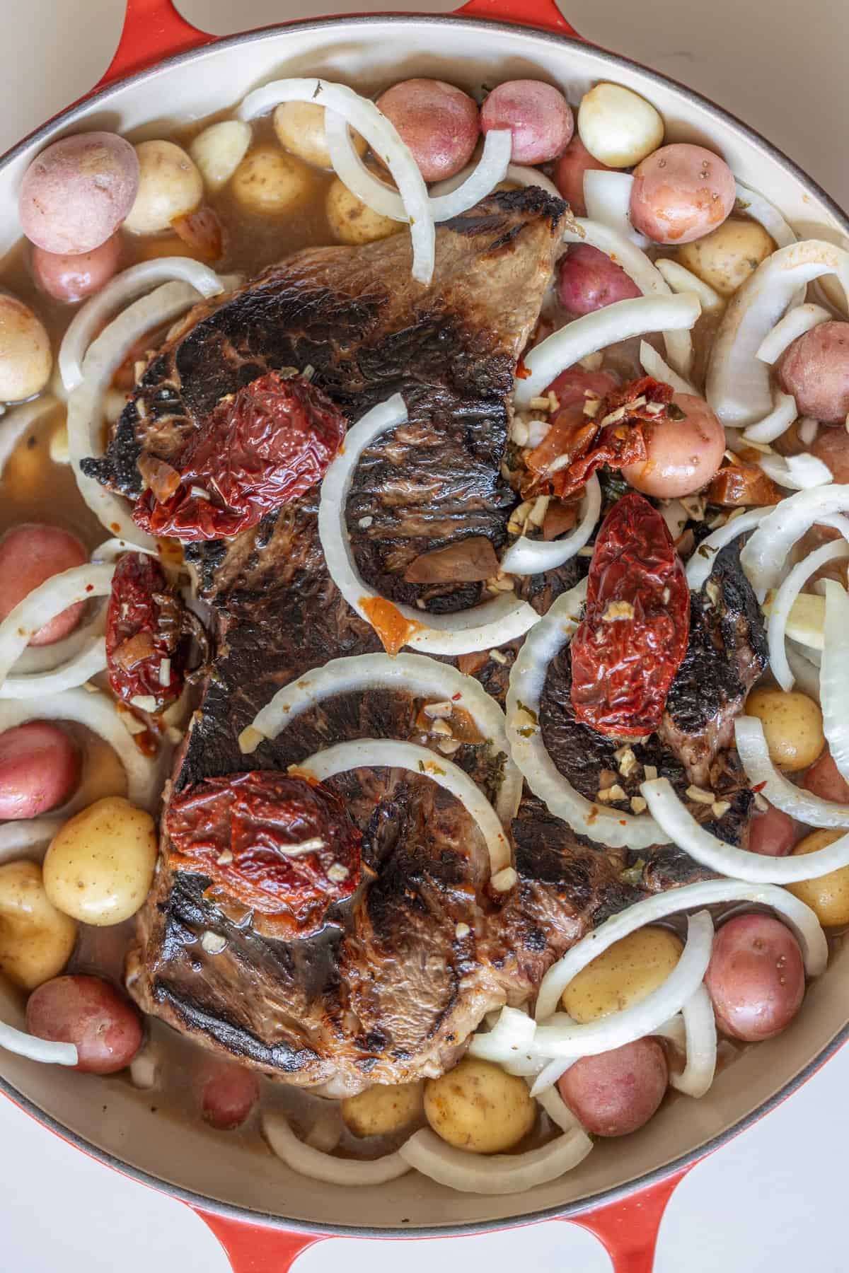A pot with seared beef, red and yellow baby potatoes, sliced onions, whole garlic cloves, and sun-dried tomatoes, ready to be cooked.