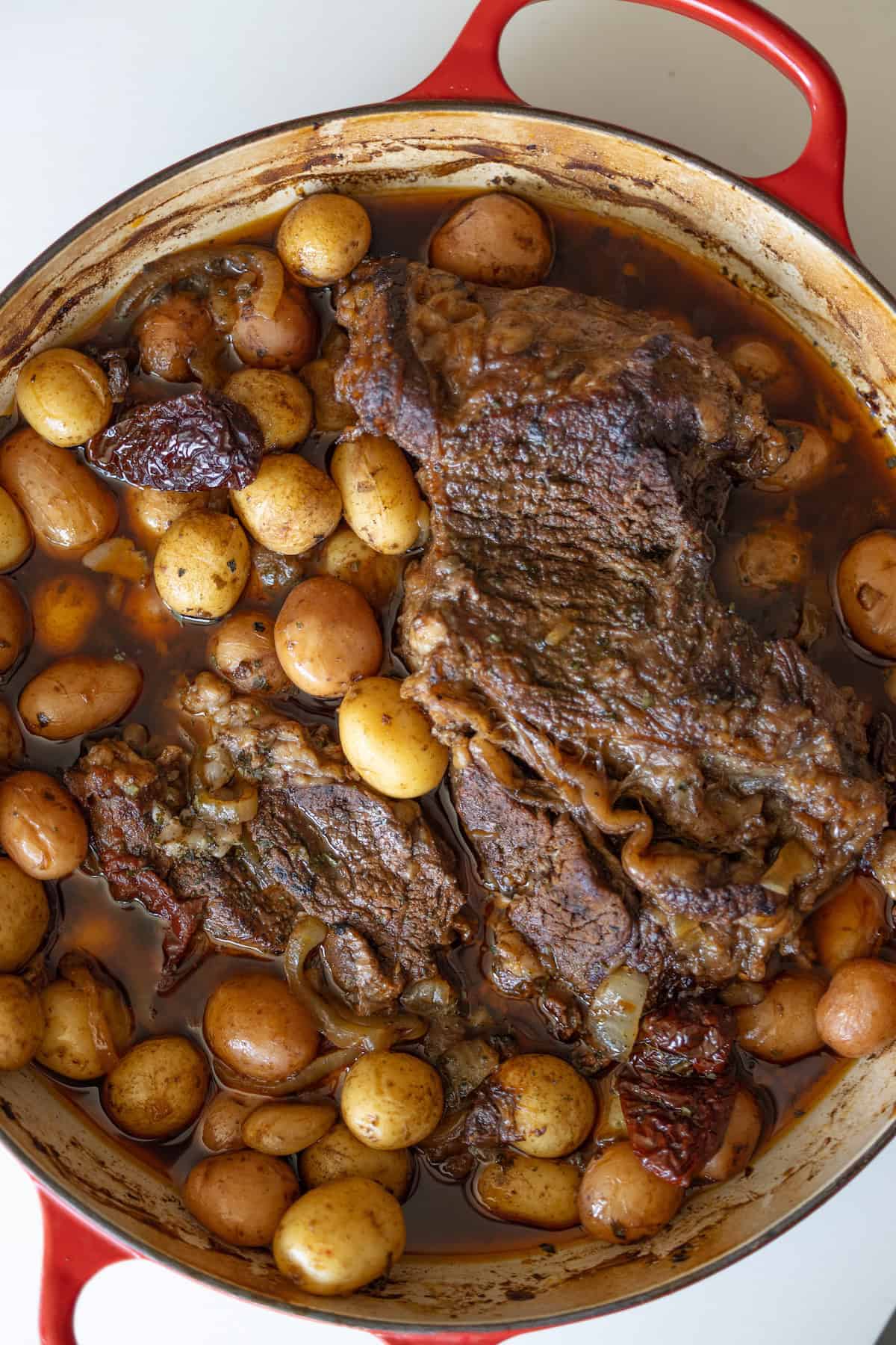 A red pot filled with cooked pot roast, baby potatoes, onions, and sun-dried tomatoes in a brown broth.
