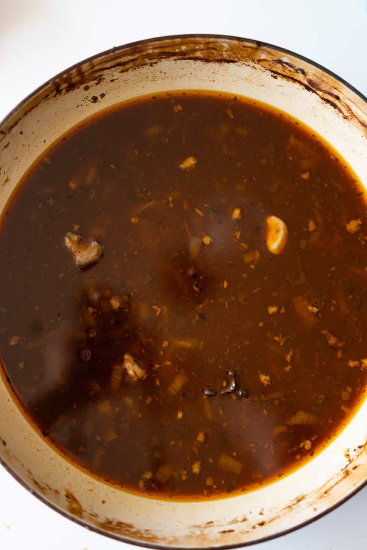 A pot filled with dark brown broth, containing pieces of meat, herbs, and visible chunks of garlic.