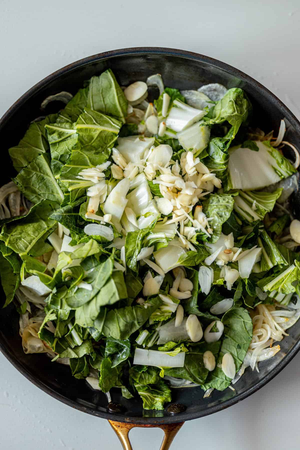 A frying pan filled with chopped bok choy, sliced garlic, slivered almonds, and pieces of fried potsticker, all ready to be cooked.