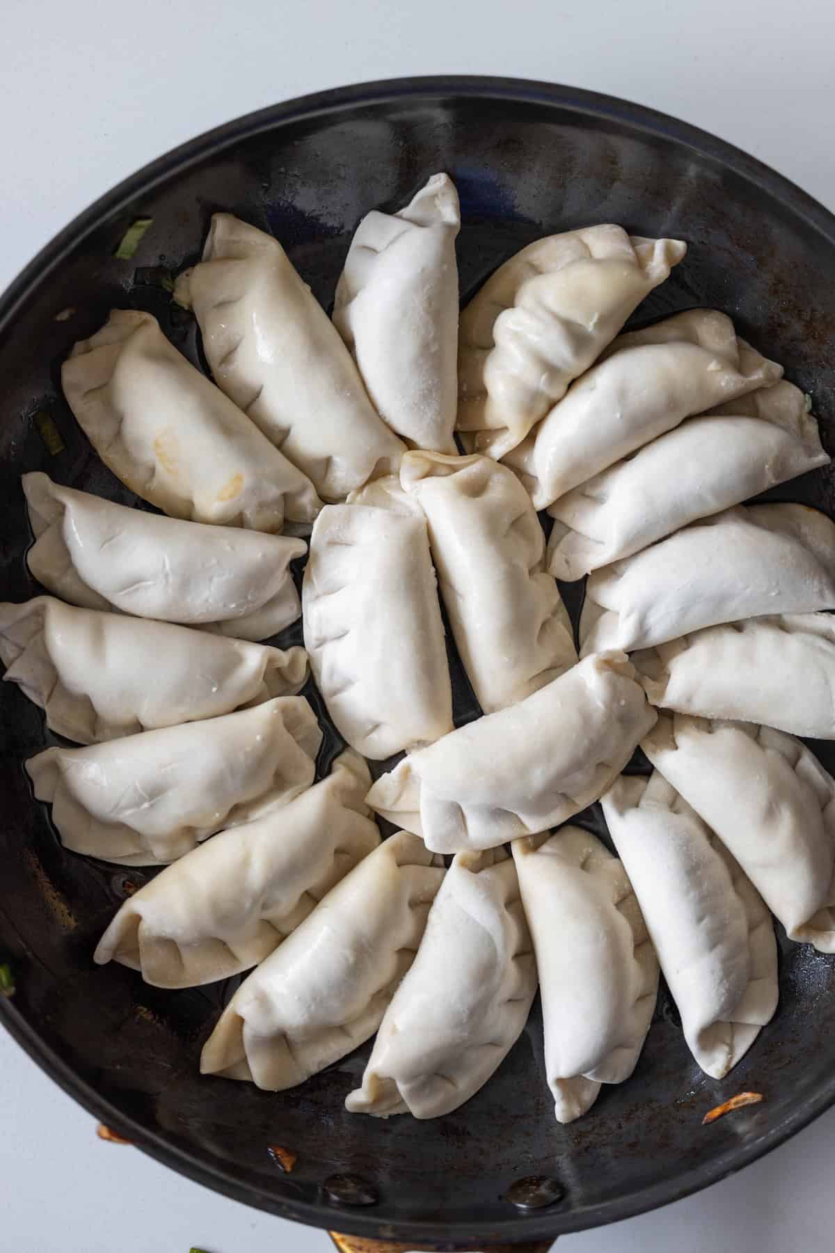 A frying pan filled with uncooked dumplings, some with crisp Bok Choy tucked inside, arranged in a circular pattern and ready to be cooked into golden fried potstickers.