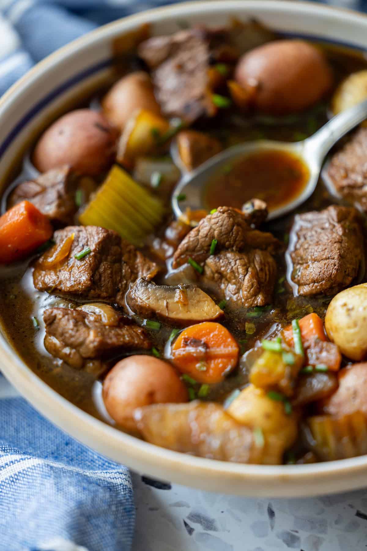 A bowl of beef stew with chunks of beef, potatoes, carrots, celery, and a spoon resting in the broth.