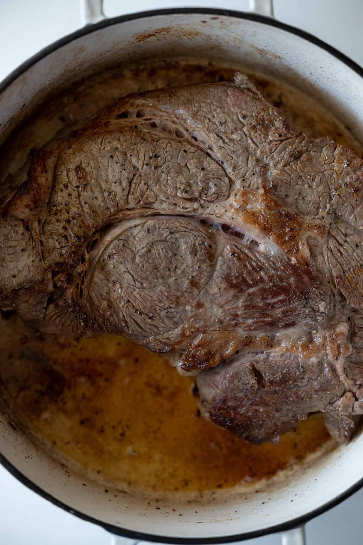 A large piece of seared beef roast browning in a white enameled cast iron pot.