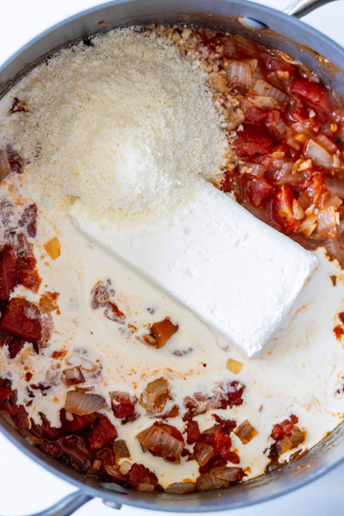 A pot containing cooked onions, diced tomatoes, a block of cream cheese, grated Parmesan cheese, cream, and zucchini—this Marry Me Zucchini is a creamy vegetarian recipe ready to be mixed together.