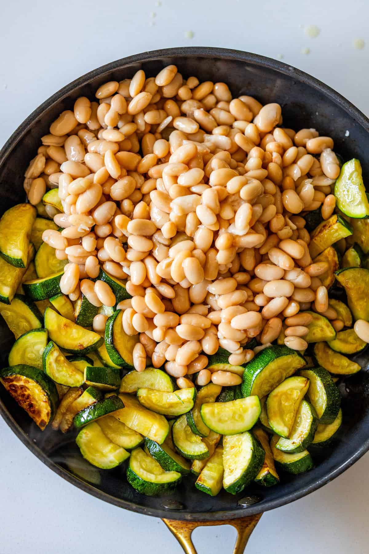 A skillet filled with cooked sliced zucchini and a pile of white beans on top, ready to be mixed together for a dish that will make you say "marry me" with every bite.